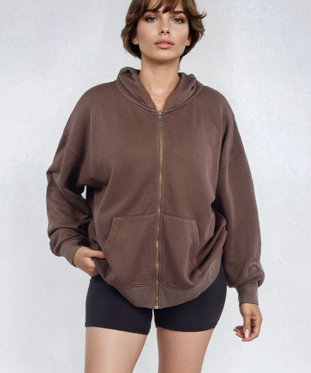 Woman wearing a relaxed brown full zip French Terry hoodie and black shorts on a white background
