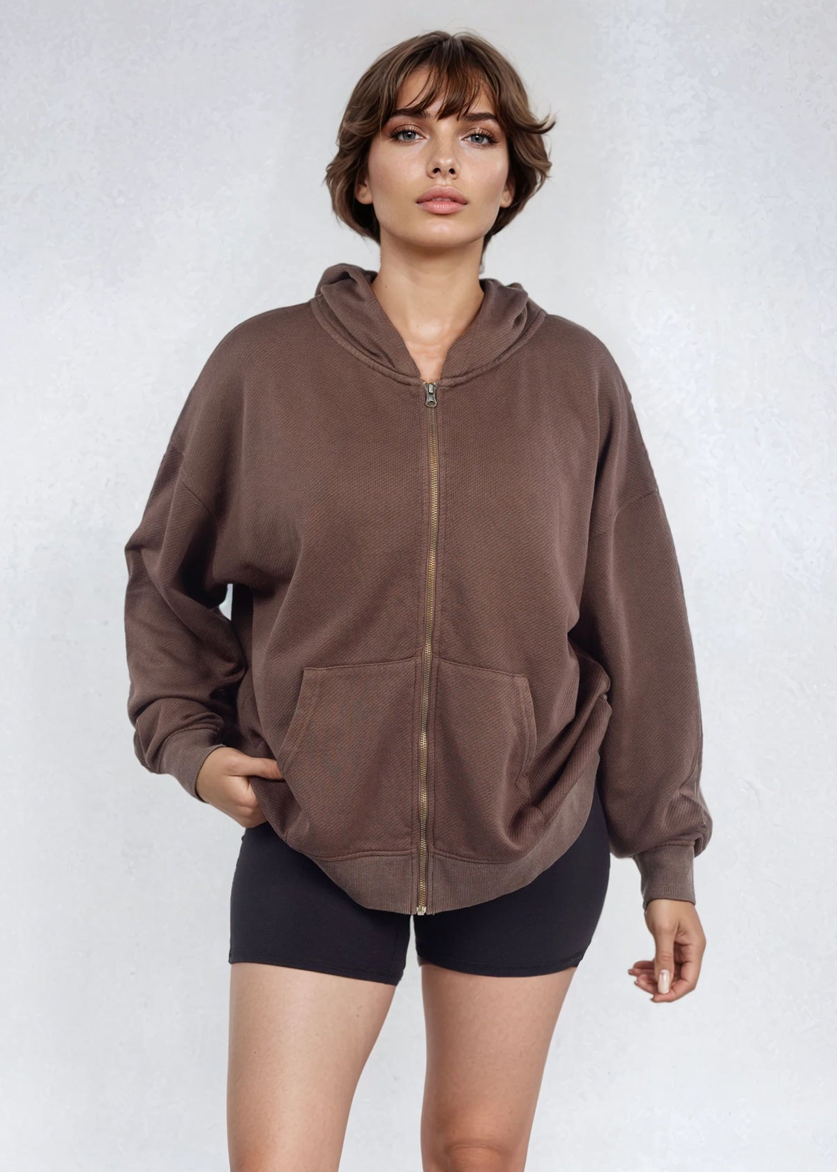 Woman wearing a relaxed brown full zip French Terry hoodie and black shorts on a white background